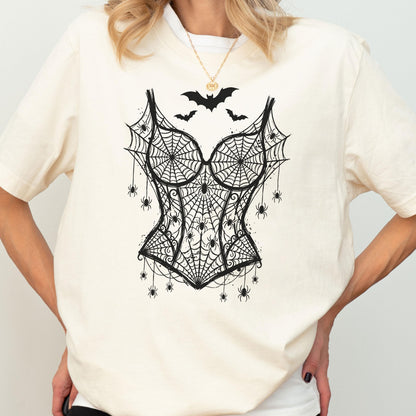 May include: A cream-colored short-sleeve t-shirt featuring a black graphic print of a corset made of spiderwebs. The design includes multiple small spiders hanging from the webbed corset and three black bats flying above the neckline.