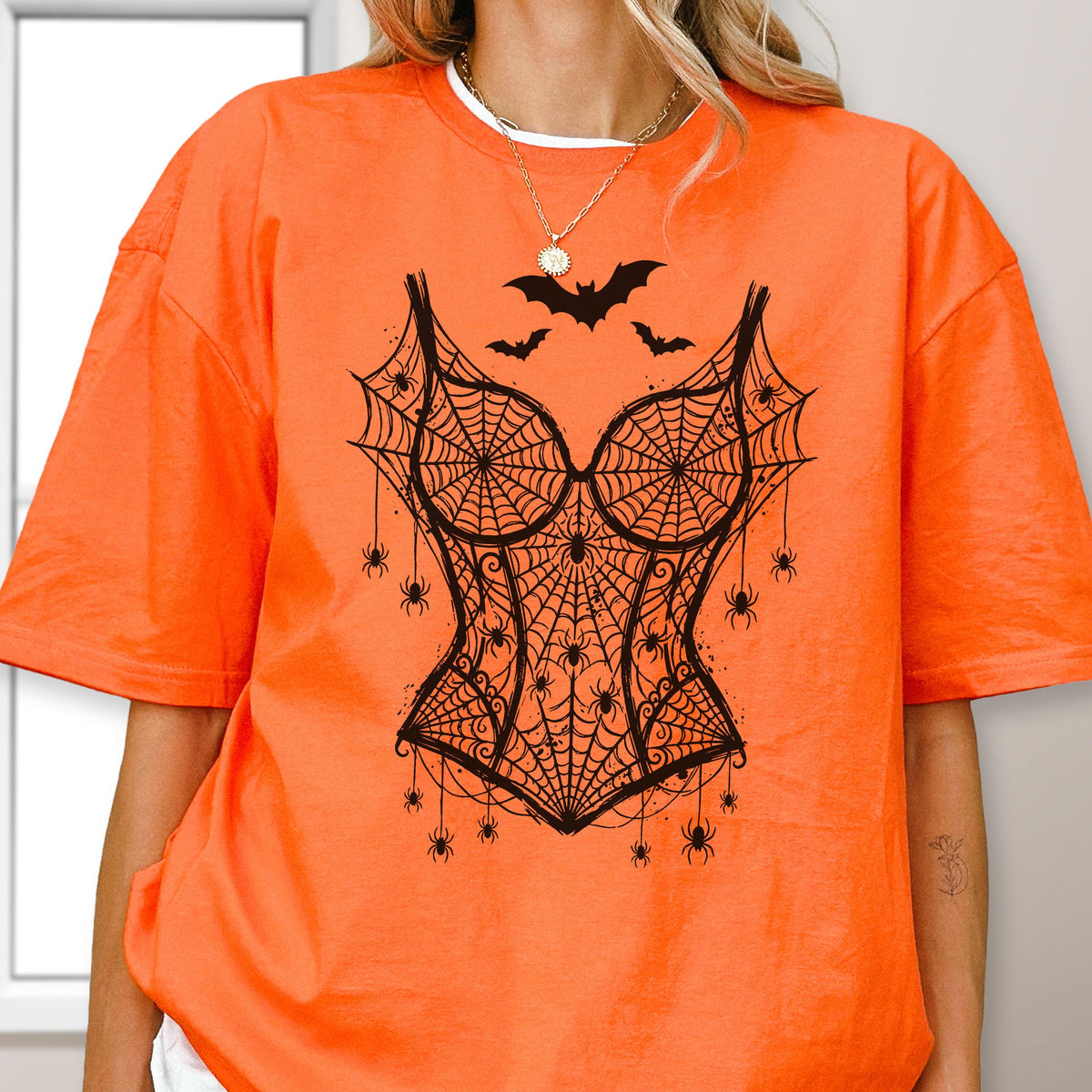 May include: A bright orange short-sleeved t-shirt featuring a black graphic print of a corset design made of spiderwebs. The corset illustration includes multiple small spiders hanging from the web pattern and three black bats flying above it.