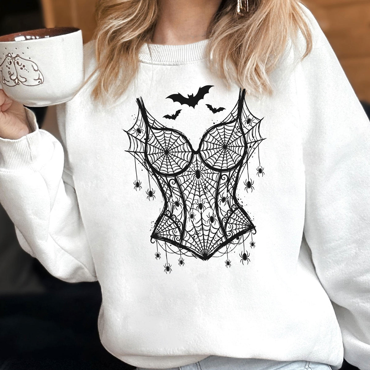 May include: A white long-sleeve sweatshirt featuring a black graphic print of a corset made of spiderwebs, adorned with multiple small spiders and three bats flying above the design. The fabric is plain white with a ribbed crew neckline.