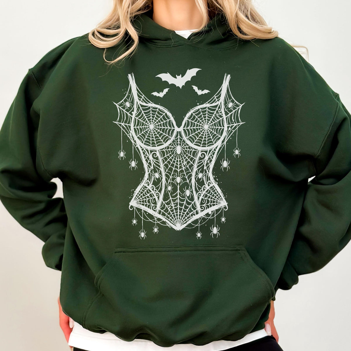 May include: A dark forest green hooded sweatshirt featuring a white graphic print on the chest. The design depicts a corset made of spiderwebs, adorned with several small spiders hanging from the web lines and two bats flying above the corset.