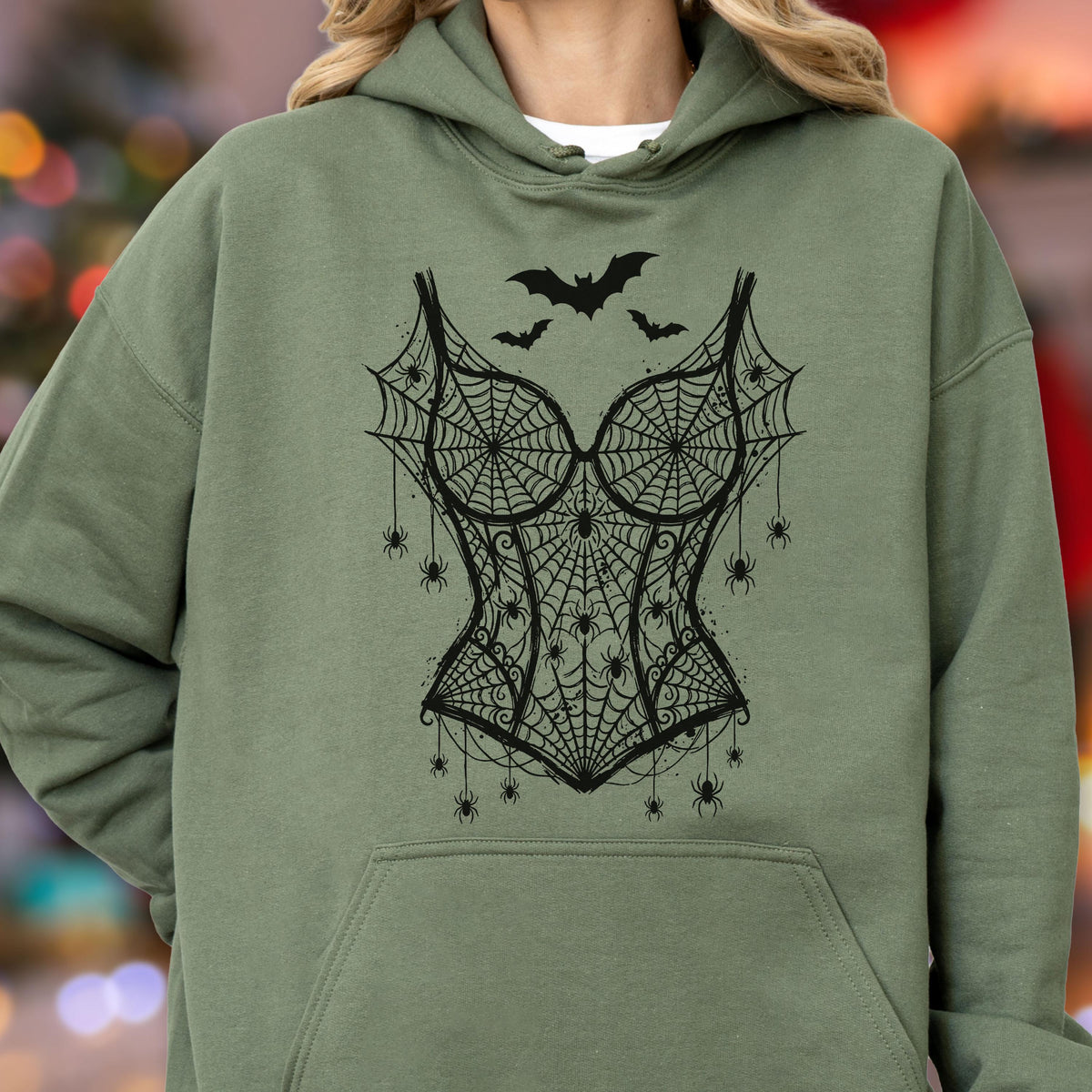 May include: An olive green hooded sweatshirt featuring a black graphic print of a corset design made of spiderwebs and spiders. The design includes three black bats flying above the corset, with multiple small spiders hanging from the webbed fabric.
