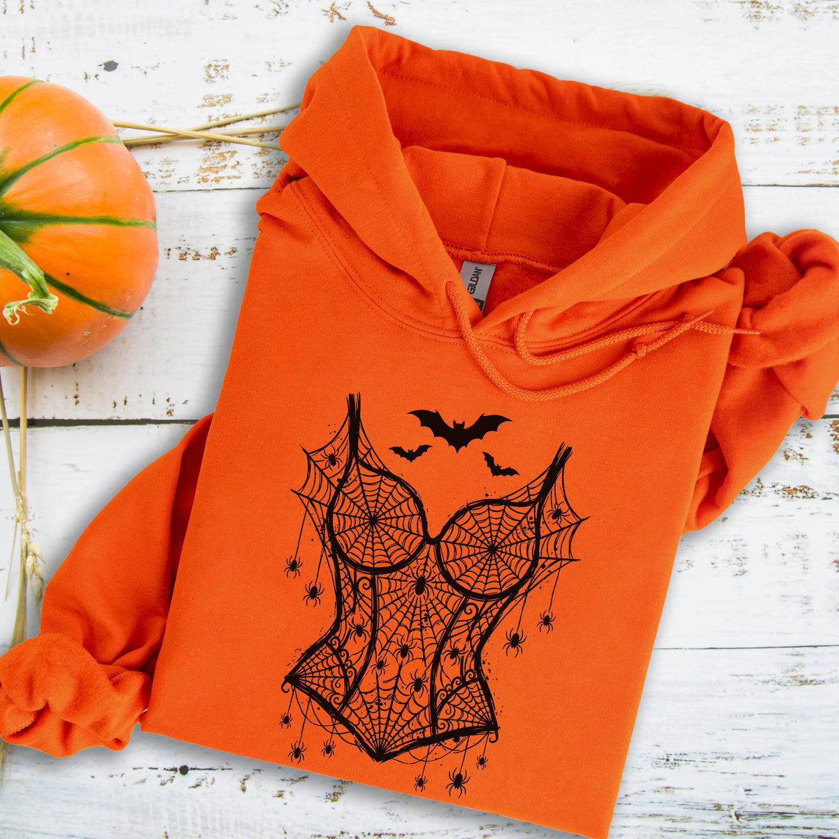 May include: A bright orange hooded sweatshirt featuring a black graphic print on the front. The design depicts a corset shape formed by spiderwebs, accented with multiple small spiders and three flying bats positioned above the corset.
