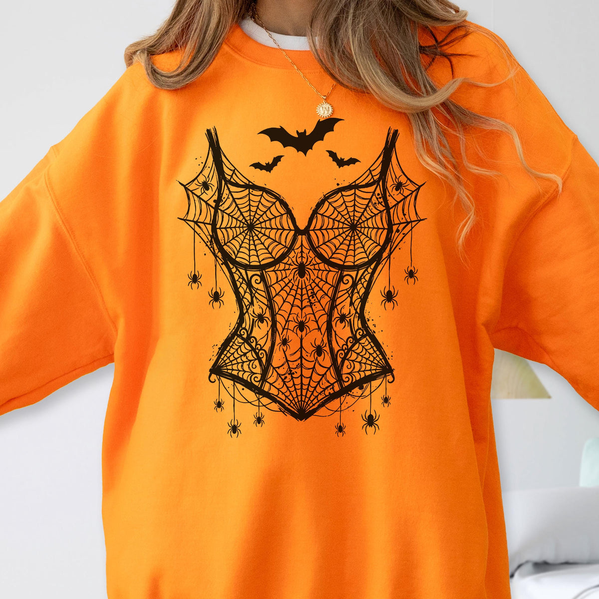 May include: A bright orange long-sleeve sweatshirt featuring a black graphic print of a corset design made of spiderwebs. The design includes multiple small spiders hanging from the web pattern and three bats flying above the corset shape.