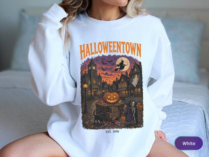 May include: A white crewneck sweatshirt featuring a graphic print with the orange text HALLOWEENTOWN above a detailed illustration of a spooky town square at night with a large pumpkin, a witch on a broomstick, and the text EST. 1998 below.