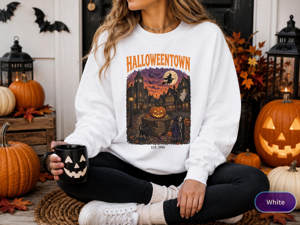 May include: A white crewneck sweatshirt featuring a graphic print of a town scene with the text HALLOWEENTOWN and EST. 1998. The illustration depicts a central pumpkin, a witch on a broomstick, a ghost, and a black cat in a dark, festive setting.