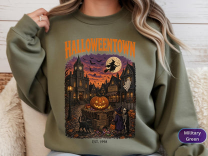 May include: A military green sweatshirt featuring a graphic print with the text HALLOWEENTOWN and EST. 1998. The illustration depicts a spooky town scene at night with a large jack-o'-lantern, a flying witch, a ghost, a black cat, and two children.