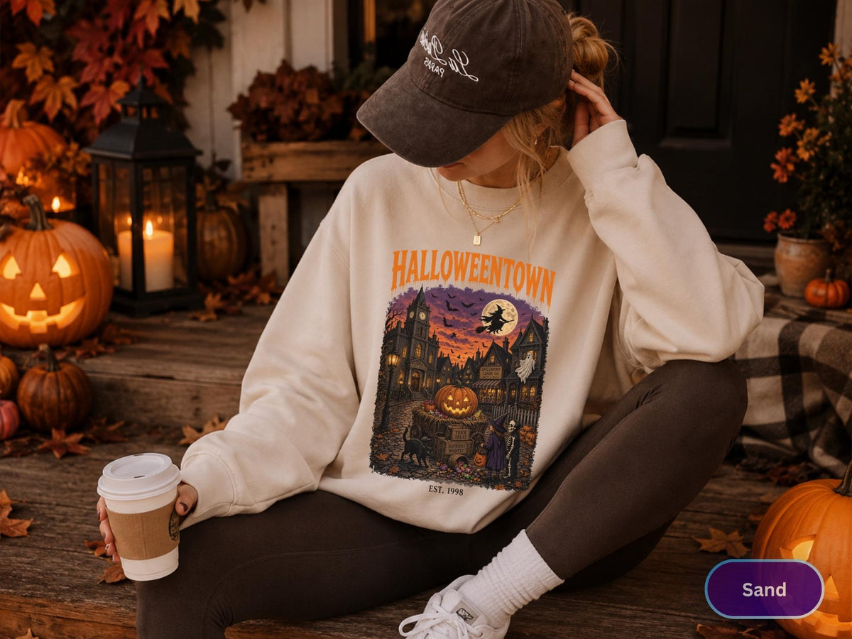 May include: A sand-colored crewneck sweatshirt featuring a graphic print with the text HALLOWEENTOWN EST. 1998. The illustration depicts a spooky town scene at sunset with a large jack-o'-lantern, a flying witch, a ghost, and figures.