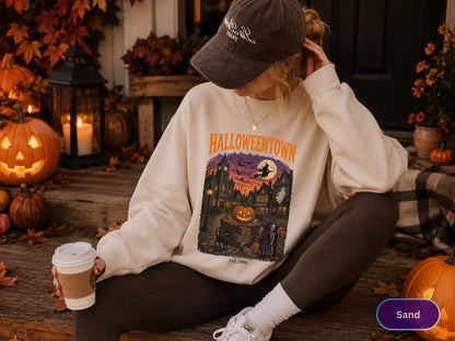 May include: A sand-colored crewneck sweatshirt featuring a graphic print with the text HALLOWEENTOWN EST. 1998. The illustration depicts a spooky town scene at sunset with a large jack-o'-lantern, a flying witch, a ghost, and figures.