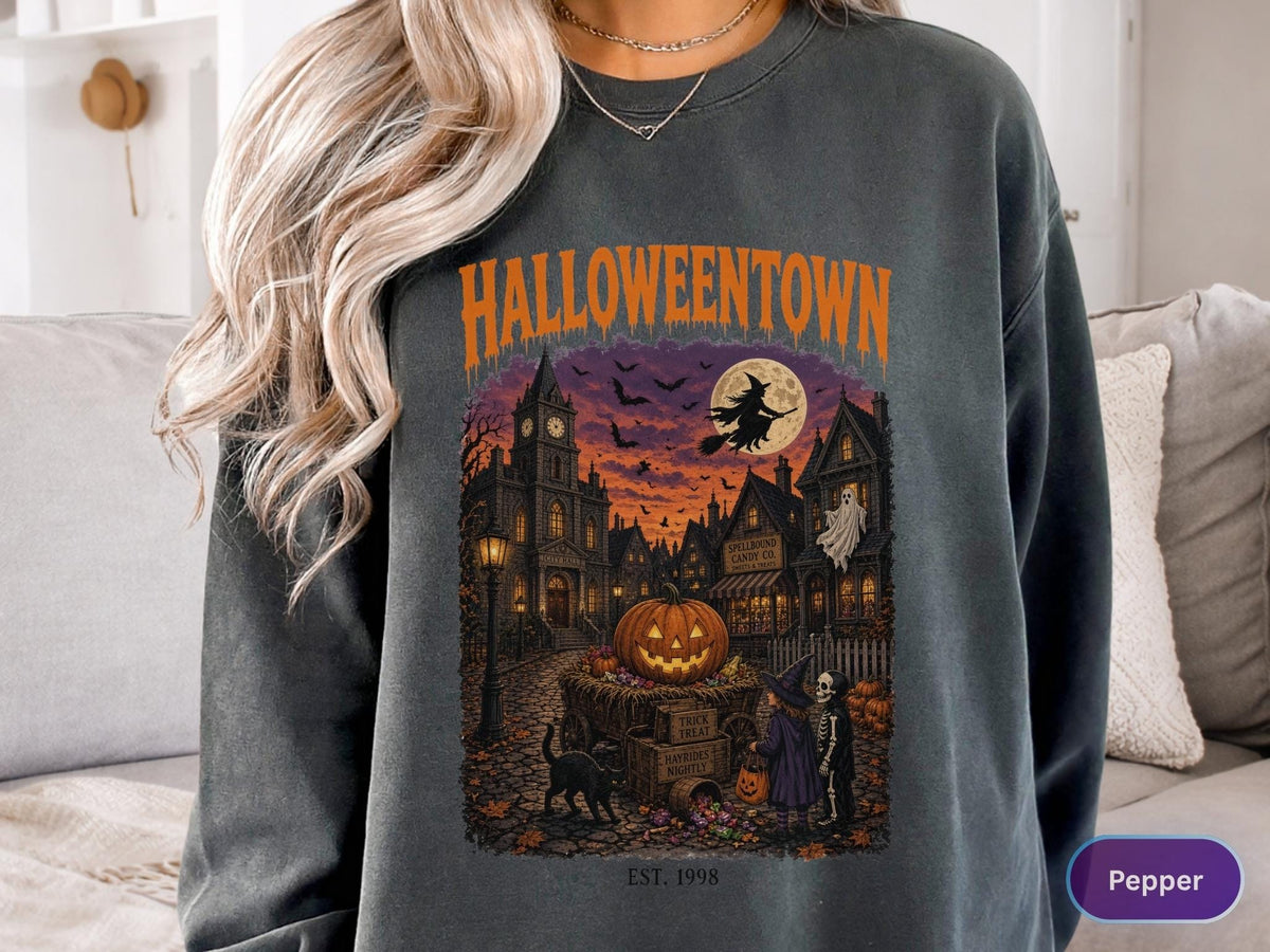 May include: A dark gray crewneck sweatshirt featuring a graphic print with the text HALLOWEENTOWN and EST. 1998. The illustration depicts a spooky town scene at night with a large jack-o'-lantern, a witch on a broomstick, a ghost, and children.