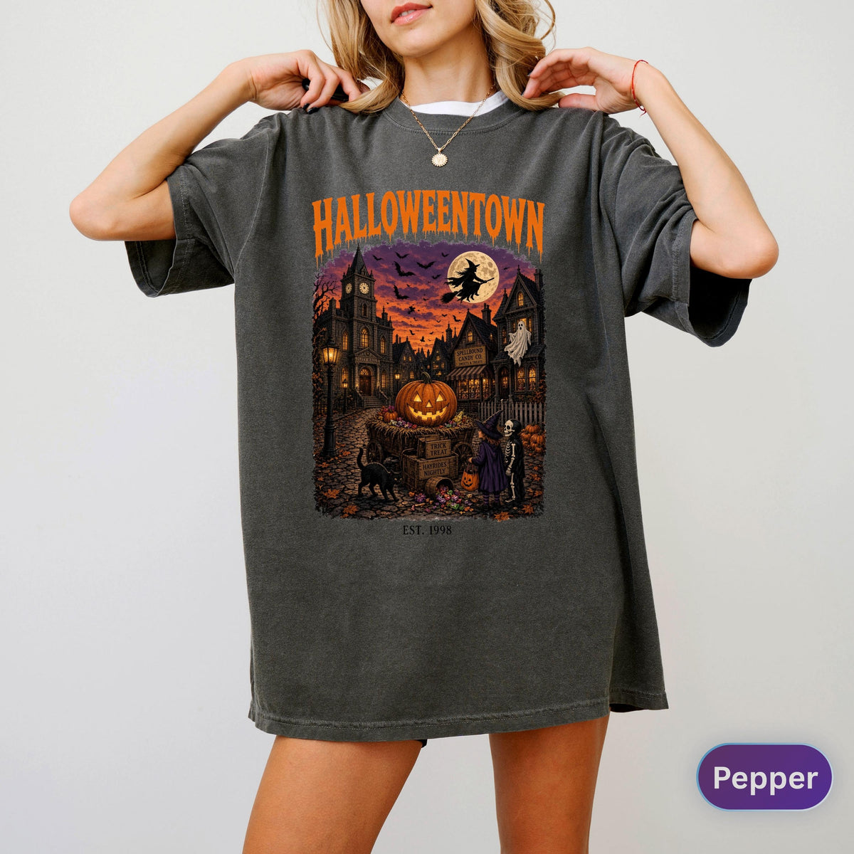 May include: A charcoal gray oversized t-shirt featuring a graphic print with the text HALLOWEENTOWN and EST. 1998. The illustration depicts a spooky town scene at night with a large orange pumpkin, a witch on a broomstick, a black cat, and a skeleton.