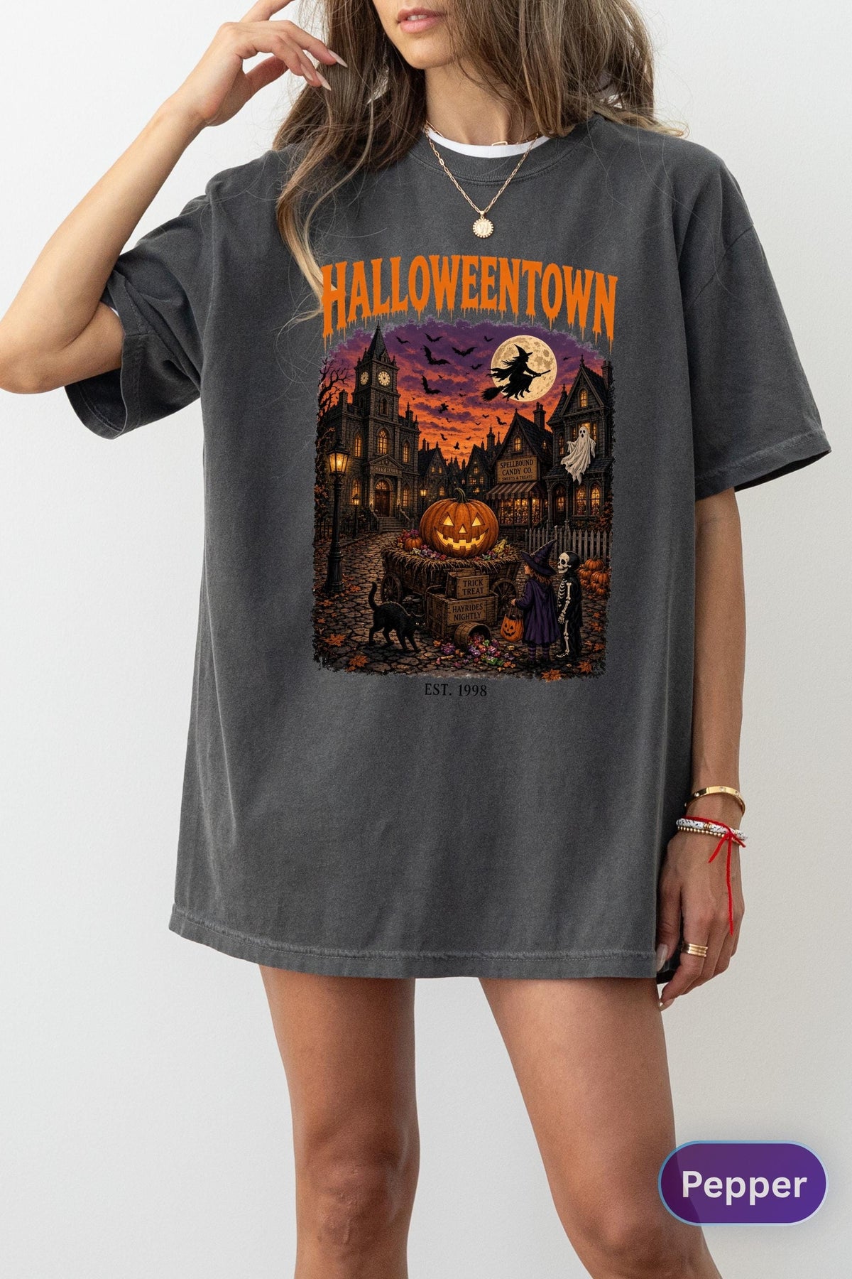 May include: A charcoal gray t-shirt featuring a graphic print with the text HALLOWEENTOWN and EST. 1998. The illustration depicts a night scene with a large jack-o'-lantern, a witch on a broomstick, a ghost, a black cat, and children in costumes.