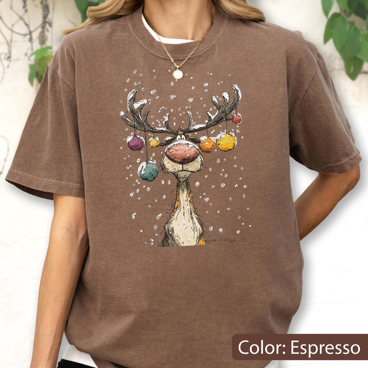 May include: An espresso brown t-shirt featuring a graphic illustration of a reindeer with large antlers decorated with colorful hanging ornaments. The reindeer has a prominent pink nose and is surrounded by white falling snow particles.