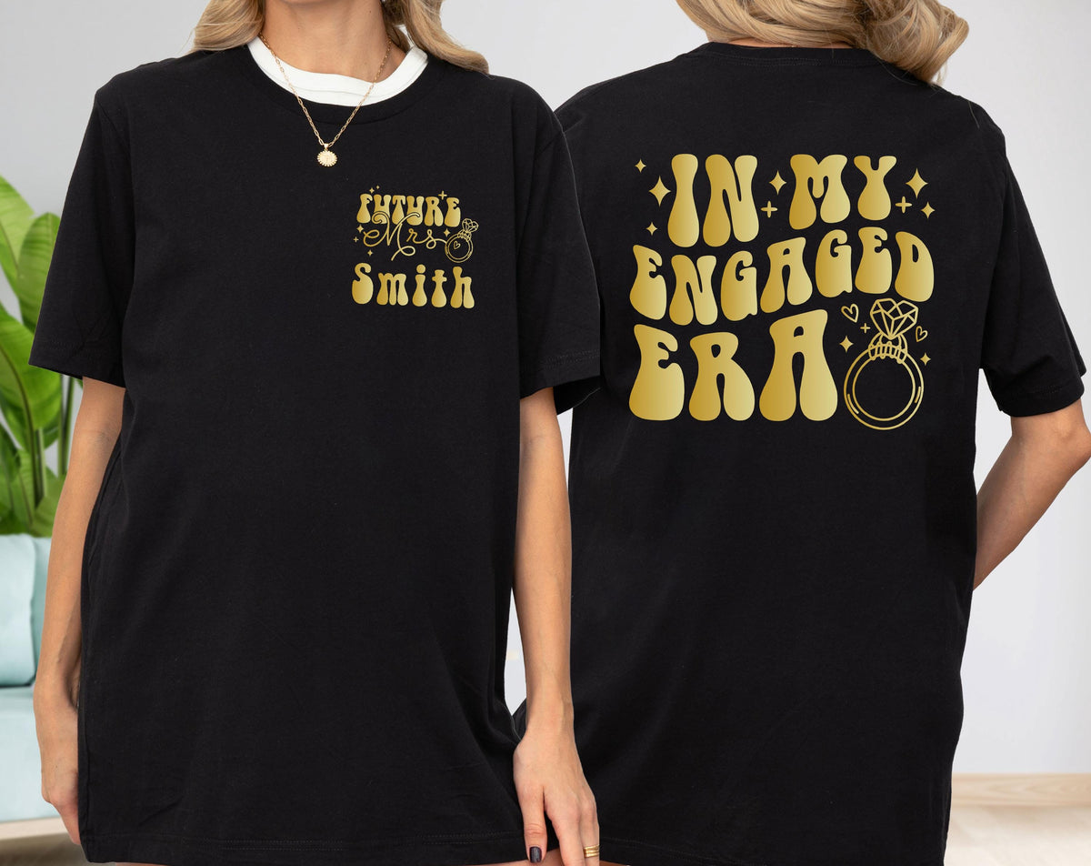 May include: A black t-shirt featuring gold text and graphics. The front left chest displays "Future Mrs. Smith" with a small ring icon. The back features the large gold text "IN MY ENGAGED ERA" accompanied by a diamond ring graphic and small stars.
