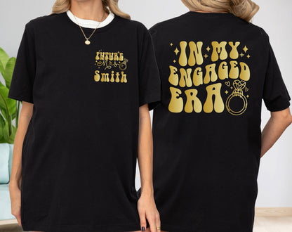 May include: A black t-shirt featuring gold text and graphics. The front left chest displays "Future Mrs. Smith" with a small ring icon. The back features the large gold text "IN MY ENGAGED ERA" accompanied by a diamond ring graphic and small stars.