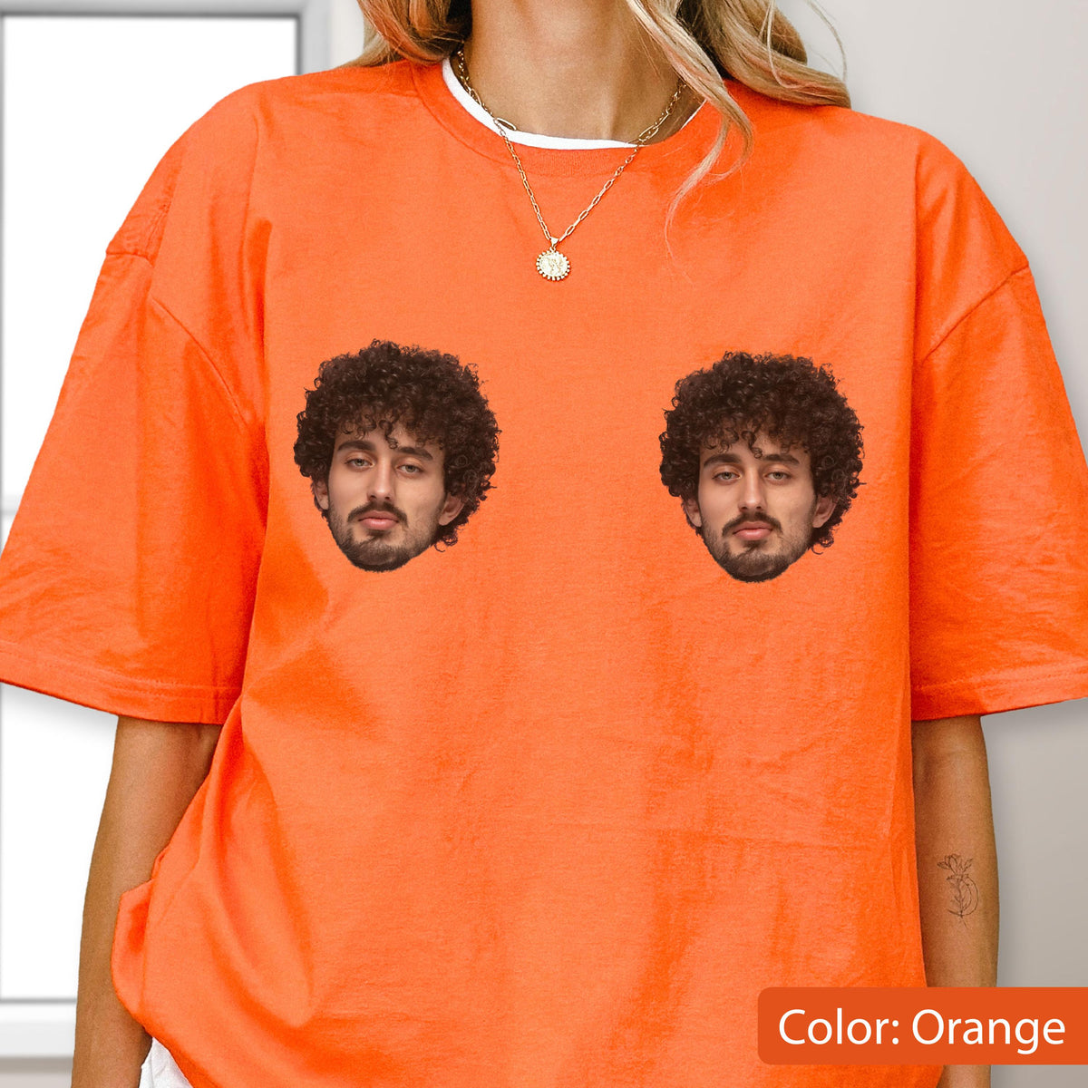 May include: A bright orange short-sleeved t-shirt featuring two identical graphic prints of a person with dark curly hair and a beard. The shirt has a crew neckline and is worn with a thin gold-toned chain necklace with a small circular pendant.