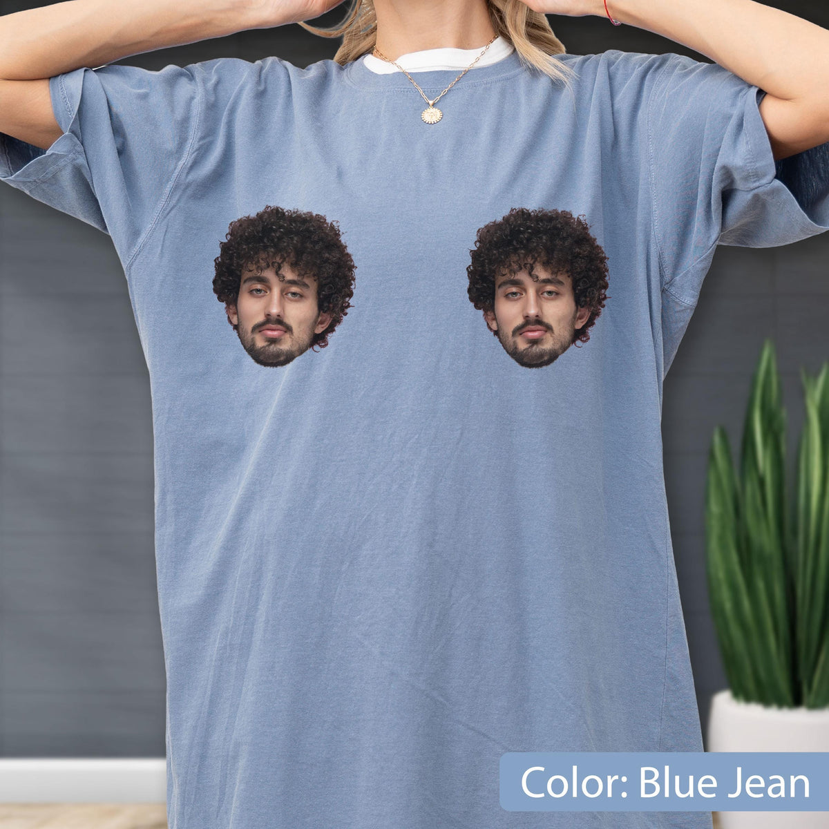 May include: A blue jean colored t-shirt featuring two identical graphic prints of a person with dark curly hair and a beard positioned on the chest area. The shirt has a crew neckline and short sleeves, showcasing a solid, muted blue fabric.