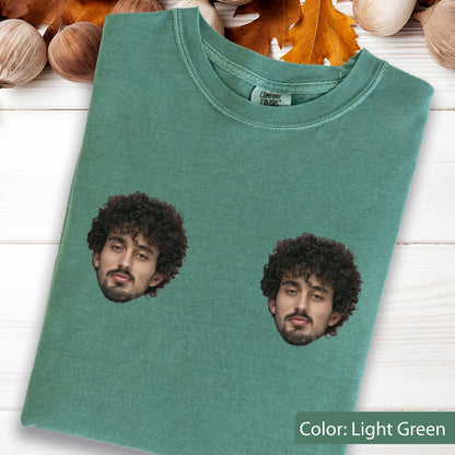 May include: A light green crew neck t-shirt featuring two identical photographic prints of a person with dark curly hair and a beard. The shirt has a Comfort Colors brand label visible at the inner neckline and a short-sleeved design.