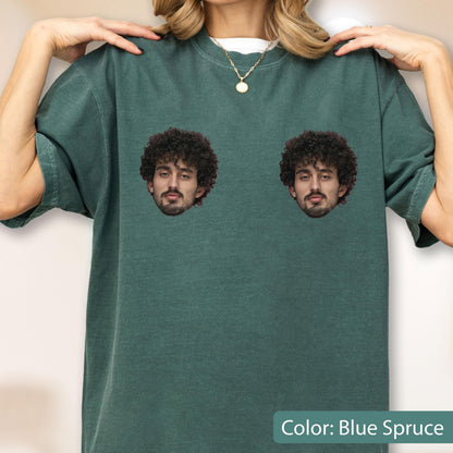 May include: A blue spruce short-sleeve t-shirt featuring two identical graphic prints of a person with dark curly hair and a beard positioned on the chest area. The shirt has a crew neckline and a relaxed fit, showcasing a solid dark teal color.