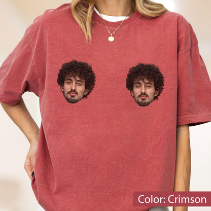 May include: A crimson, short-sleeved, crew-neck t-shirt featuring two identical graphic prints of a person with dark, curly hair and a beard positioned on the chest area. The fabric has a washed, textured appearance throughout the garment.