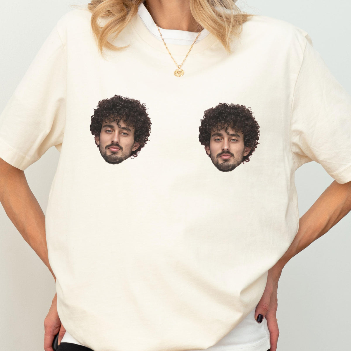May include: A cream-colored short-sleeve t-shirt featuring two identical graphic prints of a person with dark curly hair and a beard. The prints are positioned side-by-side on the chest area of the garment, which is worn with a gold chain necklace.