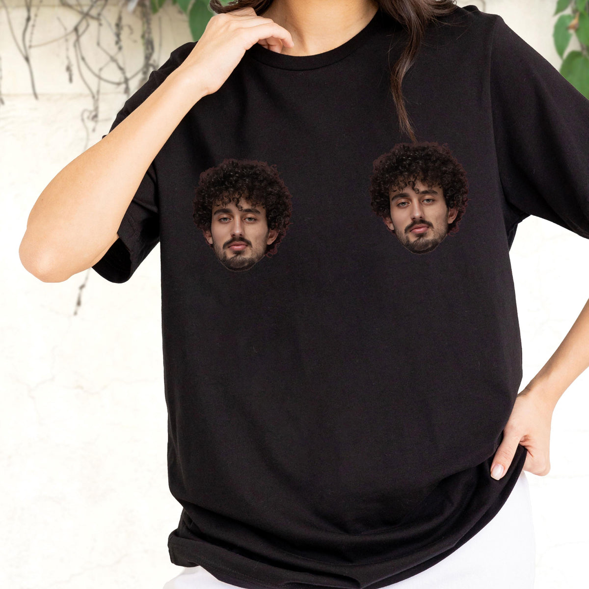 May include: A black short-sleeved t-shirt featuring two identical photographic prints of a person with dark curly hair and a beard. The two circular portraits are positioned on the chest area of the garment, which has a classic crew neckline.