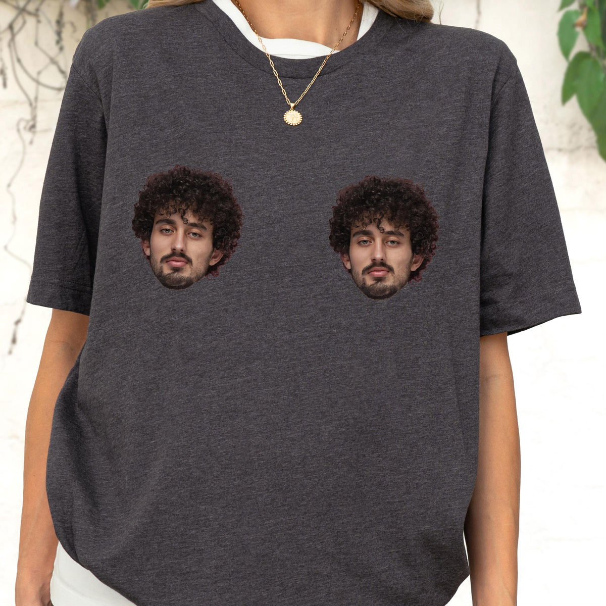 May include: A dark charcoal gray short-sleeved t-shirt featuring two identical photographic prints of a person with dark curly hair and a beard positioned on the chest area. The shirt has a crew neckline and a heathered fabric texture.