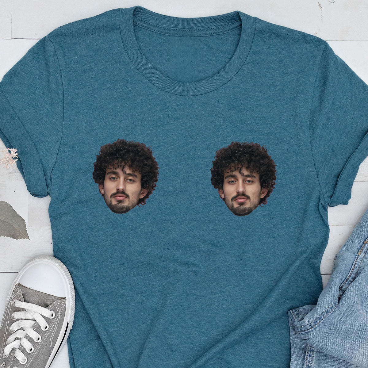 May include: A heathered teal blue short-sleeve t-shirt featuring two identical photographic prints of a person with dark curly hair and a beard positioned side-by-side on the chest area of the garment.