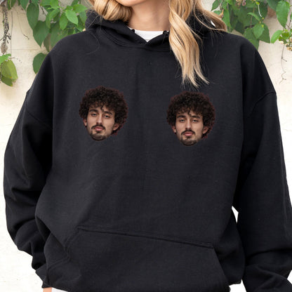 May include: A black long-sleeve hooded sweatshirt featuring two identical graphic prints of a person with dark curly hair and a beard positioned on the chest area. The garment includes a front kangaroo pocket and a drawstring hood.