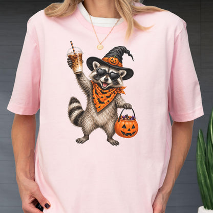 May include: A light pink t-shirt featuring a graphic of a raccoon wearing a black witch hat with a pumpkin, heart-shaped sunglasses, and an orange bandana with a bat pattern. The raccoon holds an iced coffee and a pumpkin-shaped candy bucket.