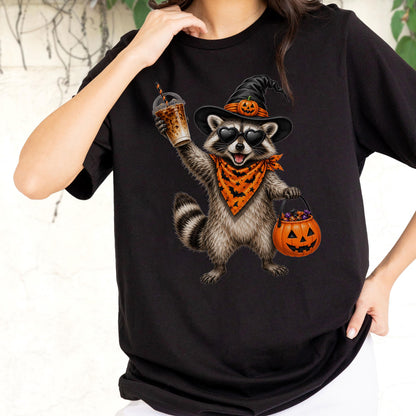 May include: A black t-shirt featuring a graphic of a raccoon wearing a black witch hat with a small pumpkin, heart-shaped sunglasses, and an orange bandana with a black bat pattern. The raccoon holds an iced coffee and a jack-o'-lantern bucket.