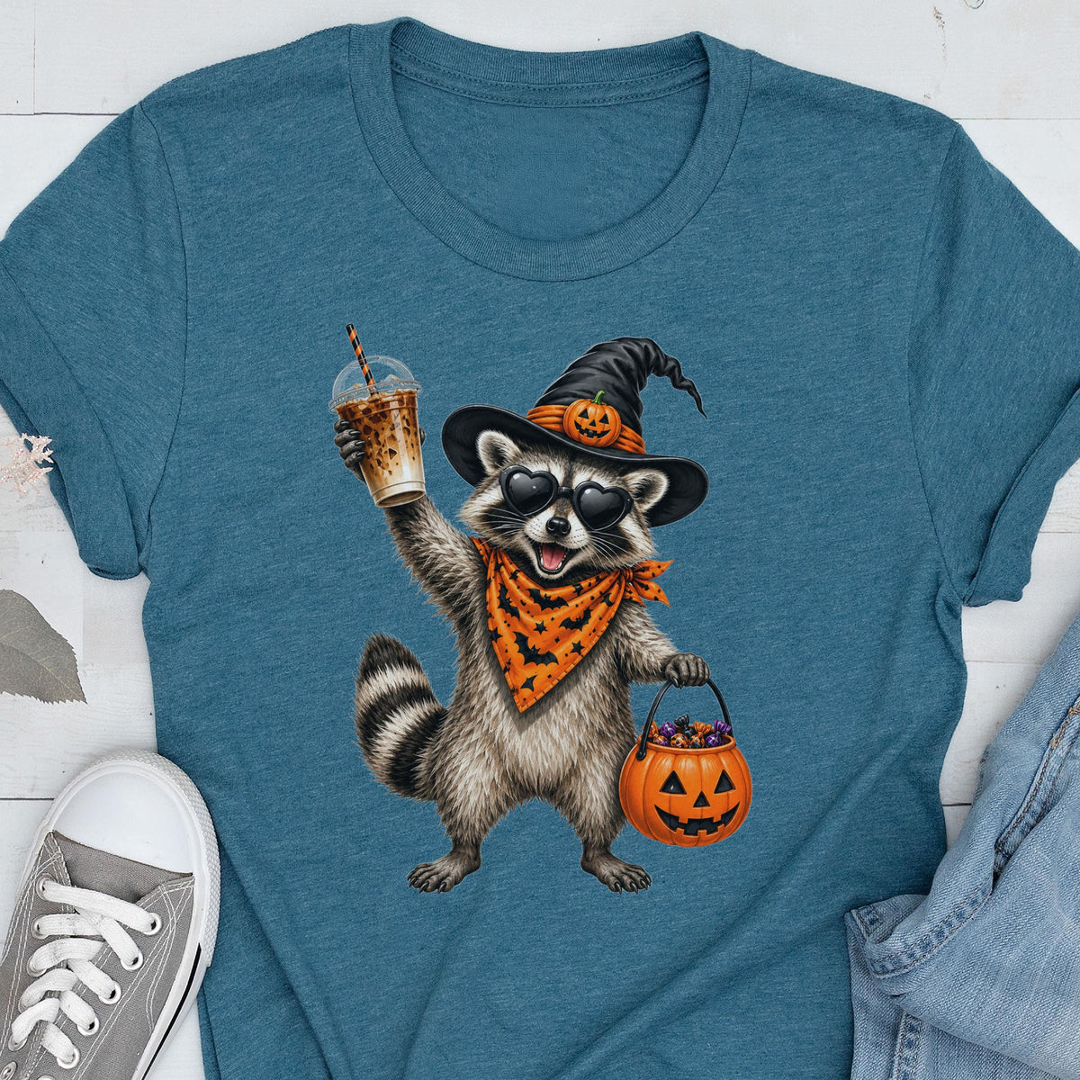 May include: A teal blue t-shirt featuring a graphic of a raccoon wearing a black witch hat with a small pumpkin, heart-shaped sunglasses, and an orange bandana with black bats. The raccoon holds an iced coffee and a jack-o'-lantern candy bucket.