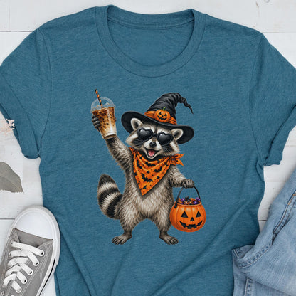 May include: A teal blue t-shirt featuring a graphic of a raccoon wearing a black witch hat with a small pumpkin, heart-shaped sunglasses, and an orange bandana with black bats. The raccoon holds an iced coffee and a jack-o'-lantern candy bucket.