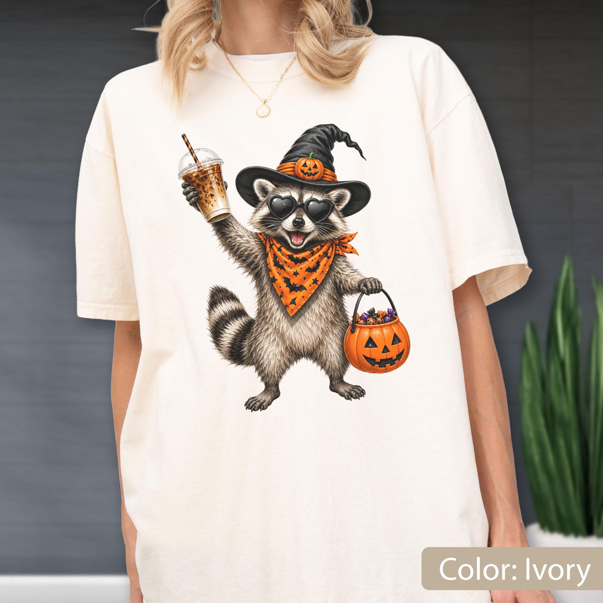 May include: An ivory t-shirt featuring a graphic of a raccoon wearing a black witch hat with a small pumpkin, heart-shaped sunglasses, and an orange bandana with a black bat pattern. The raccoon holds an iced coffee and a jack-o'-lantern bucket.