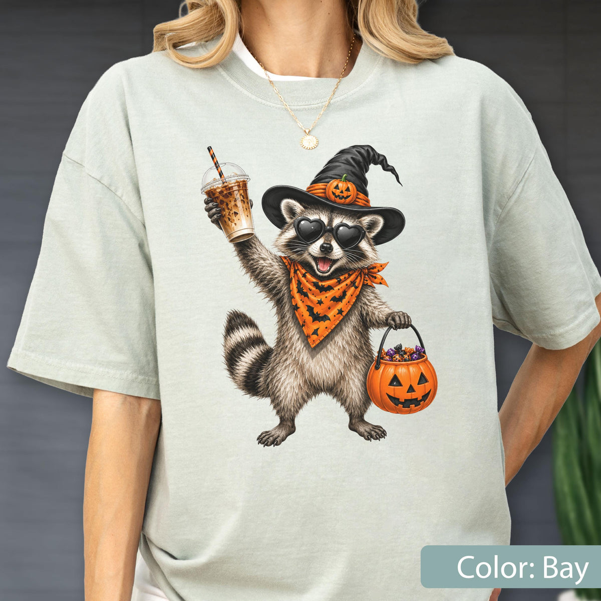 May include: A light bay-colored t-shirt featuring a graphic of a raccoon wearing a black witch hat with a small pumpkin, heart-shaped sunglasses, and an orange bandana with black bats. The raccoon holds an iced coffee and a pumpkin candy bucket.