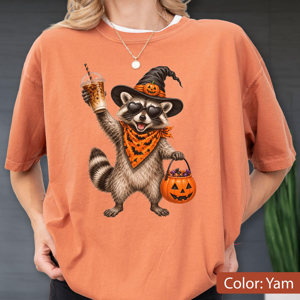 May include: An orange t-shirt featuring a graphic of a raccoon wearing a witch hat with a small pumpkin, heart-shaped sunglasses, and an orange bandana with black bats. The raccoon holds a plastic iced coffee cup and a jack-o'-lantern candy bucket.
