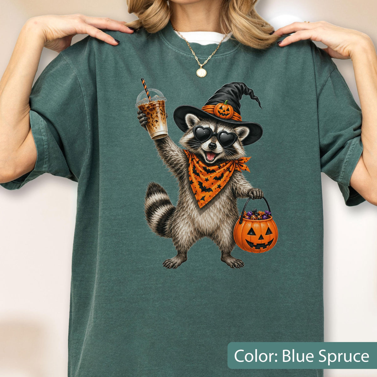 May include: A blue spruce t-shirt featuring a graphic of a raccoon wearing a witch hat with a pumpkin, heart-shaped sunglasses, and an orange bat-patterned bandana. The raccoon holds an iced coffee and a jack-o'-lantern bucket filled with candy.
