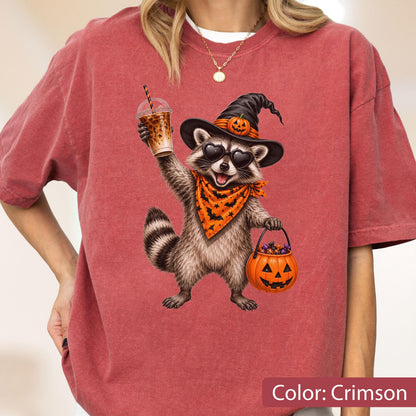 May include: A crimson short-sleeve t-shirt featuring a graphic of a raccoon wearing a black witch hat with a small pumpkin, heart-shaped sunglasses, and an orange bandana with black bats. The raccoon holds an iced coffee and a pumpkin bucket.