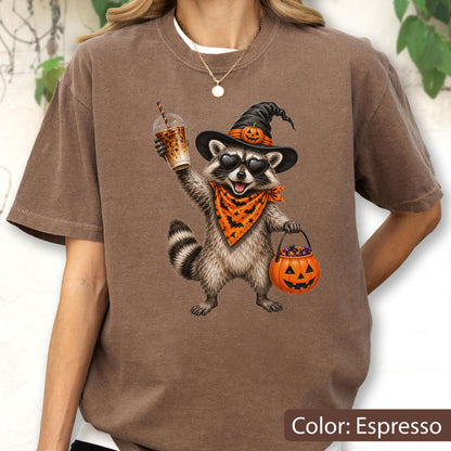 May include: An espresso-colored t-shirt featuring a graphic of a raccoon wearing a witch hat with a small pumpkin, heart-shaped sunglasses, and an orange bat-patterned bandana. The raccoon holds an iced coffee and a jack-o'-lantern candy bucket.