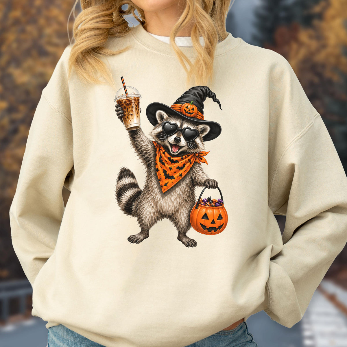 May include: A beige crewneck sweatshirt featuring a graphic of a raccoon wearing a black witch hat with a pumpkin accent, heart-shaped sunglasses, and an orange bandana. The raccoon holds a plastic iced coffee cup and a jack-o'-lantern candy bucket.