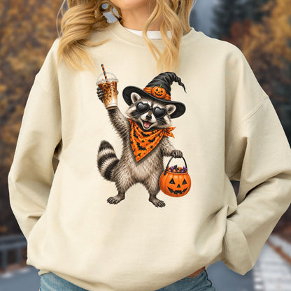 May include: A beige crewneck sweatshirt featuring a graphic of a raccoon wearing a black witch hat with a pumpkin accent, heart-shaped sunglasses, and an orange bandana. The raccoon holds a plastic iced coffee cup and a jack-o'-lantern candy bucket.