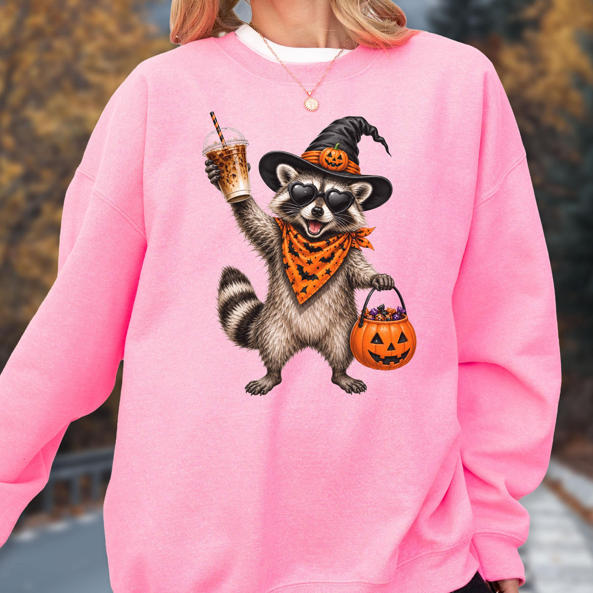 May include: A bright pink crewneck sweatshirt featuring a graphic of a raccoon wearing a black witch hat with a pumpkin, heart-shaped sunglasses, and an orange bandana with black bats. The raccoon holds an iced coffee and a pumpkin candy bucket.