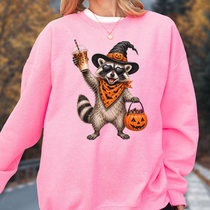 May include: A bright pink crewneck sweatshirt featuring a graphic of a raccoon wearing a black witch hat with a pumpkin, heart-shaped sunglasses, and an orange bandana with black bats. The raccoon holds an iced coffee and a pumpkin candy bucket.