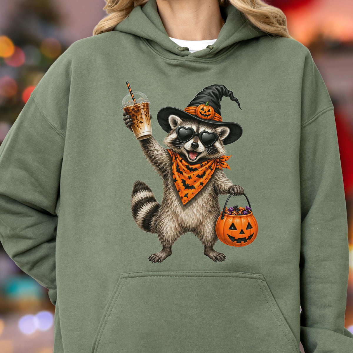 May include: An olive green hooded sweatshirt featuring a graphic of a raccoon wearing a black witch hat with a small pumpkin, heart-shaped sunglasses, and an orange bandana with black bats. The raccoon holds an iced coffee and a pumpkin bucket.