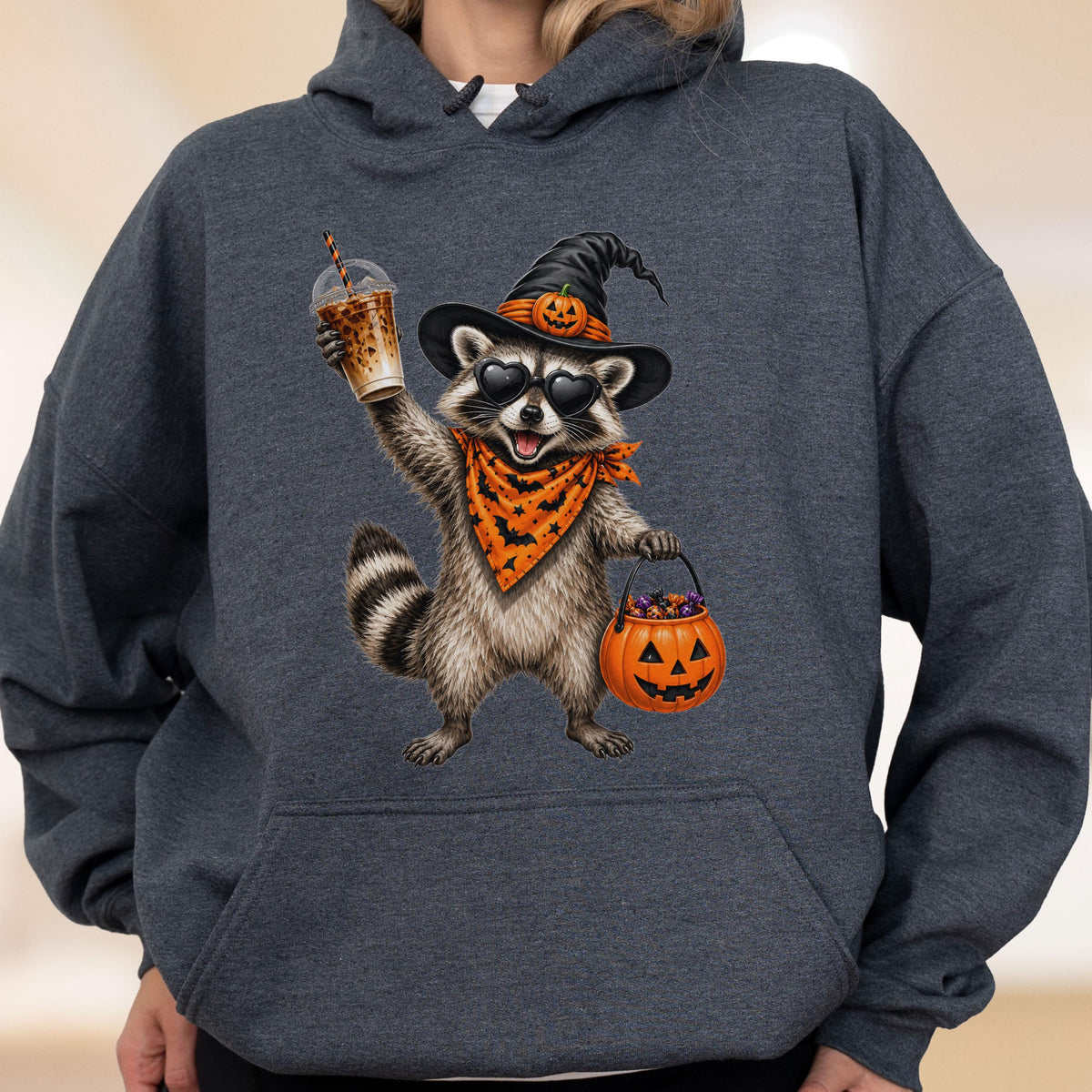 May include: A dark heather gray hooded sweatshirt featuring a graphic of a raccoon wearing a black witch hat with a small pumpkin, heart-shaped sunglasses, and an orange bandana with black bats. The raccoon holds an iced coffee and a pumpkin bucket.