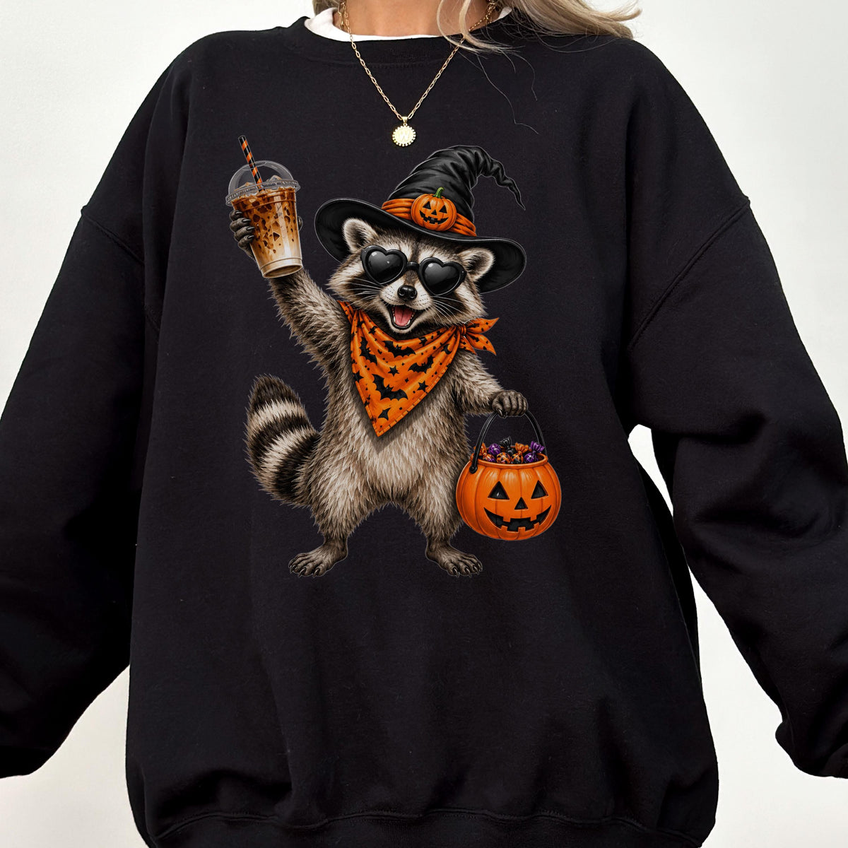 May include: A black crewneck sweatshirt featuring a graphic of a raccoon wearing a witch hat with a small pumpkin, heart-shaped sunglasses, and an orange bandana with black bats. The raccoon holds an iced coffee and a jack-o'-lantern candy bucket.