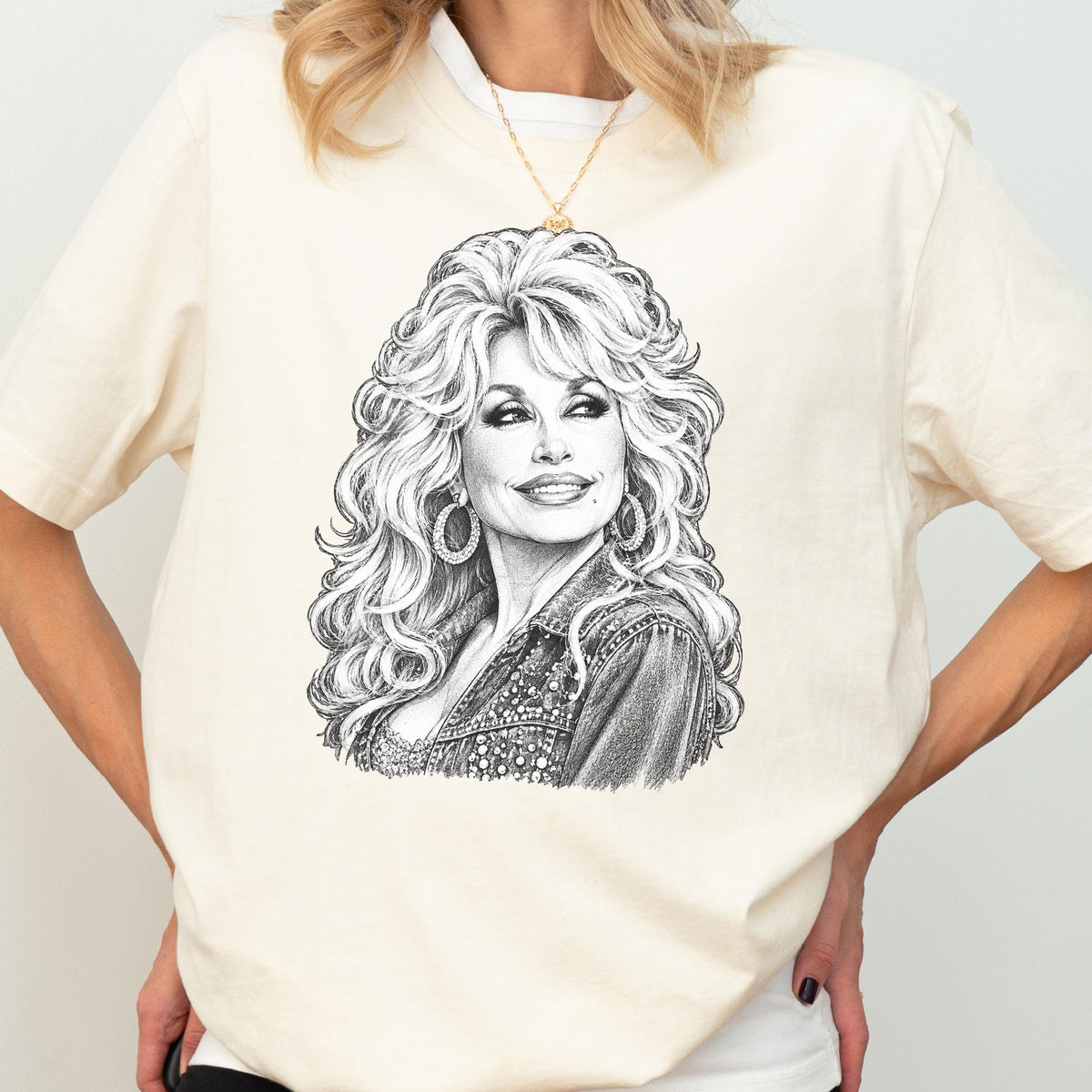 May include: A cream-colored short-sleeve t-shirt featuring a detailed black and white pencil-style sketch of a woman with voluminous blonde hair, wearing large hoop earrings and a denim jacket, centered on the front of the garment.