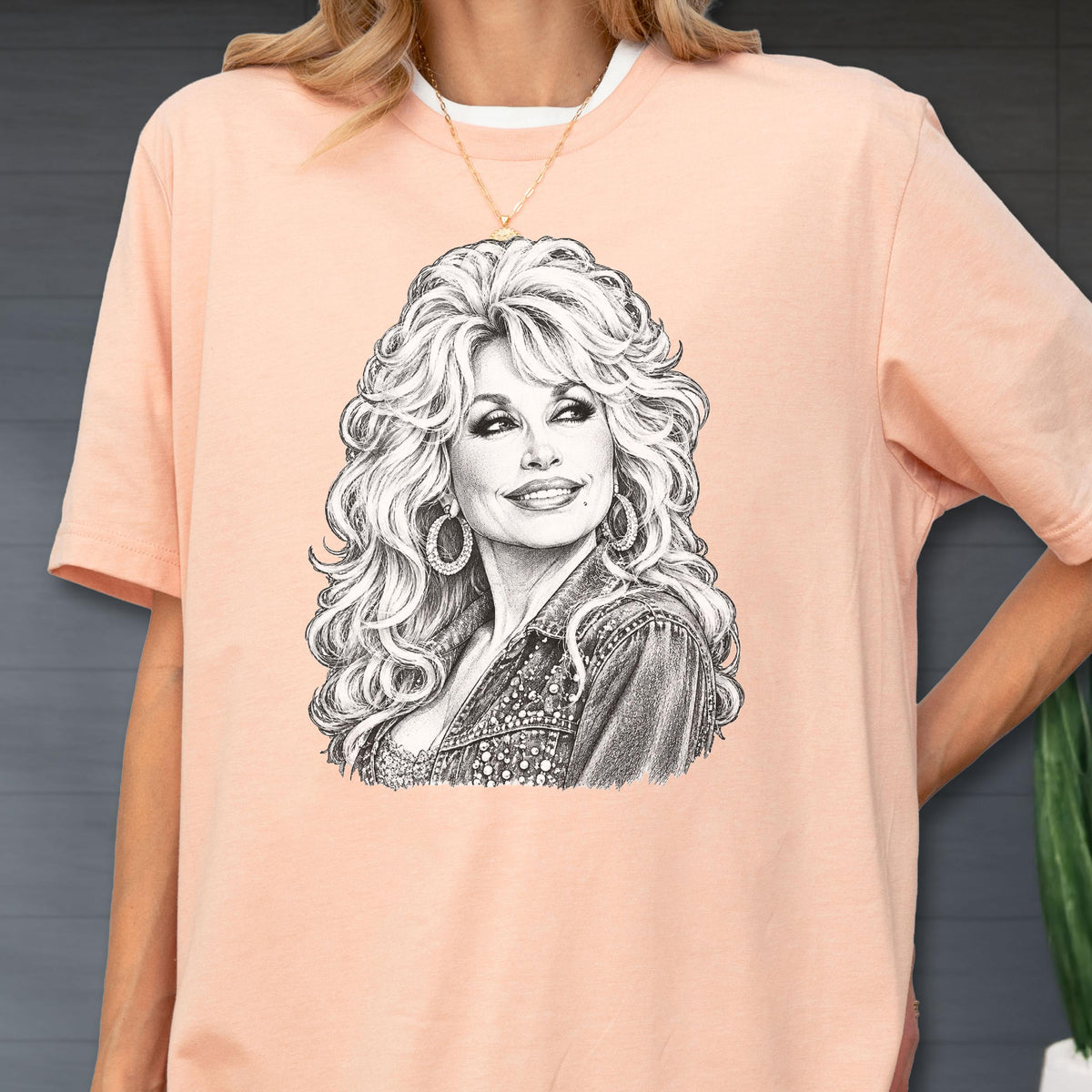May include: A peach-colored short-sleeve t-shirt featuring a detailed black and white pencil-style sketch graphic of a woman with voluminous wavy hair, wearing large hoop earrings and a textured jacket, centered on the front of the garment.