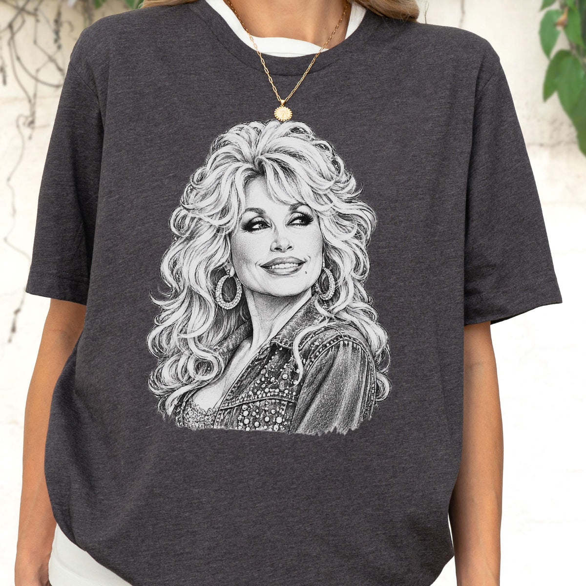 May include: A charcoal gray short-sleeve t-shirt featuring a detailed white sketch illustration of a woman with voluminous blonde hair, wearing large hoop earrings and a denim jacket. The graphic is centered on the chest of the garment.