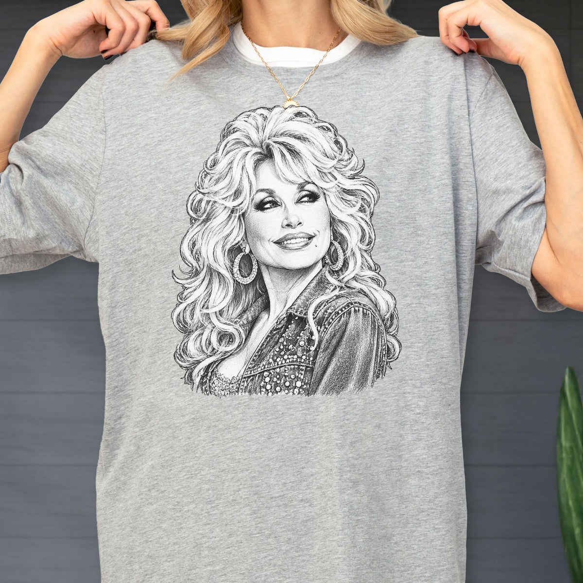 May include: A heather gray short-sleeve t-shirt featuring a detailed black and white pencil-style sketch of a woman with voluminous blonde hair, wearing hoop earrings and a textured jacket, centered on the front of the garment.