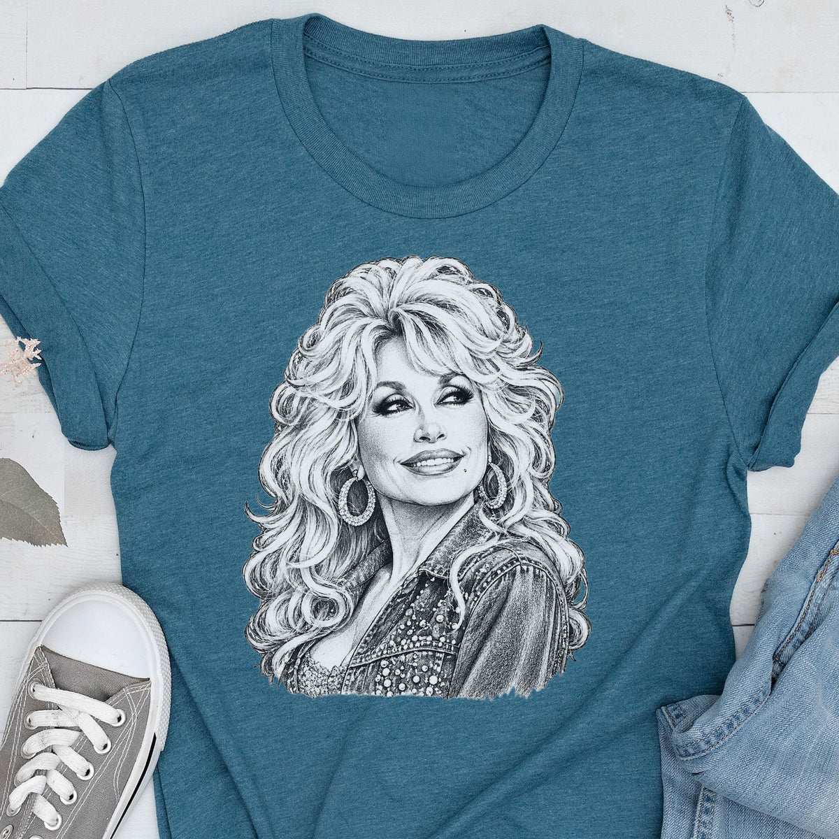 May include: A teal blue short-sleeved t-shirt featuring a detailed black and white pencil-style sketch of a person with voluminous blonde hair, wearing hoop earrings and a collared jacket, centered on the front of the garment.